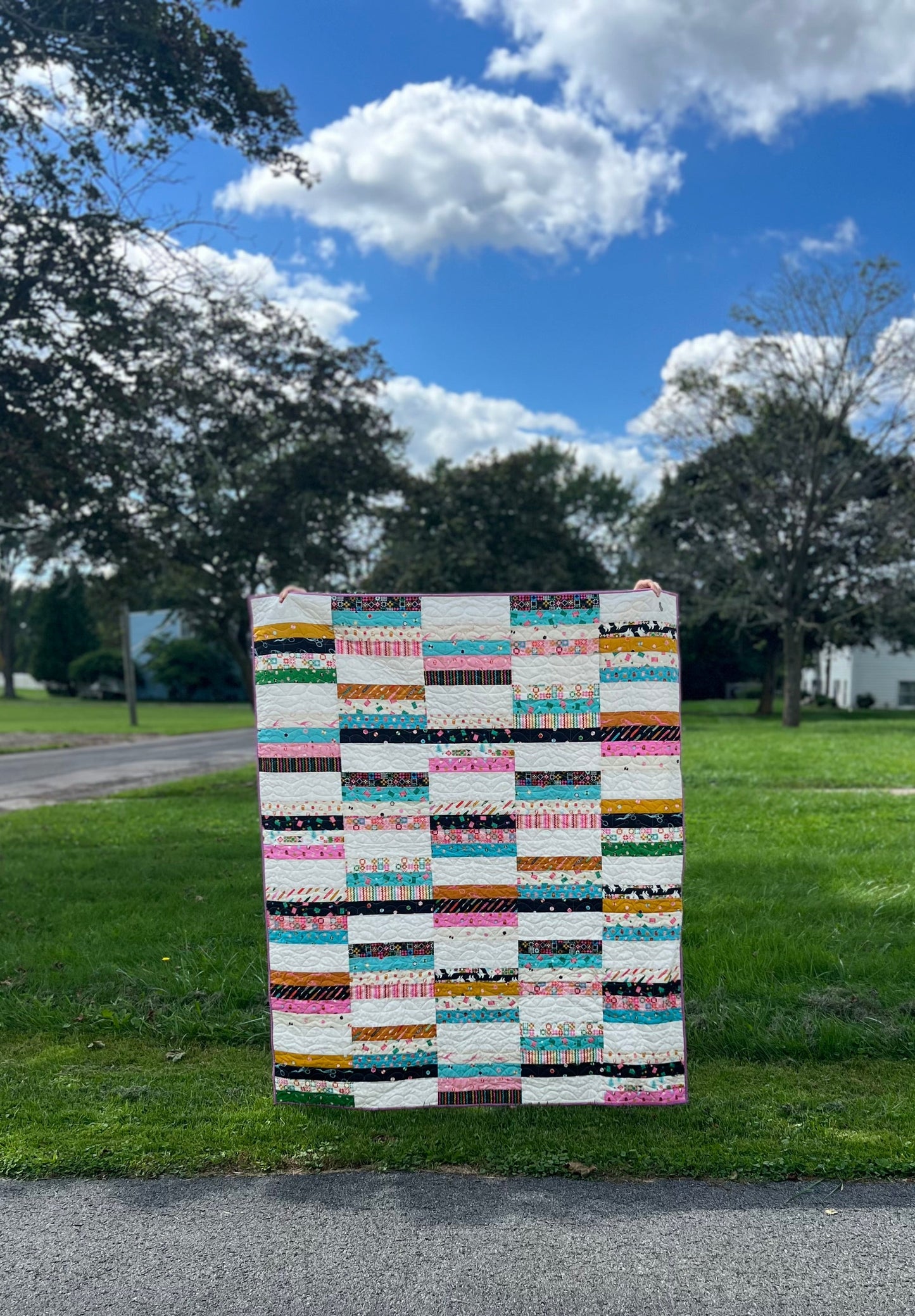Finished Hannah Quilt displayed outdoors showing colorful strip-pieced patchwork with turquoise, pink, black, gold, and white 2.5-inch strips in a simple grid layout